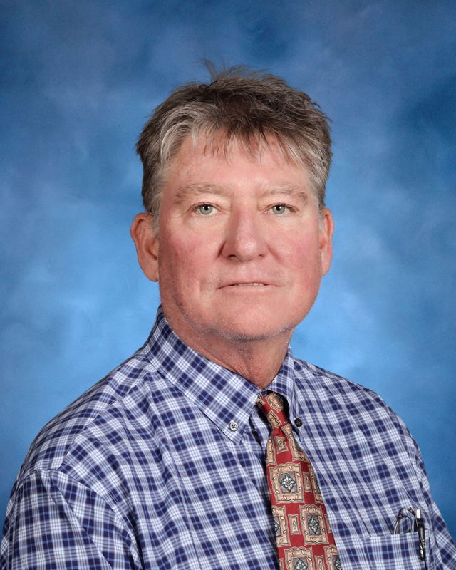A middle-aged man in a plaid shirt and tie poses against blue, reflecting the distinguished spirit of St. Paul’s School for boys on the Northshore.