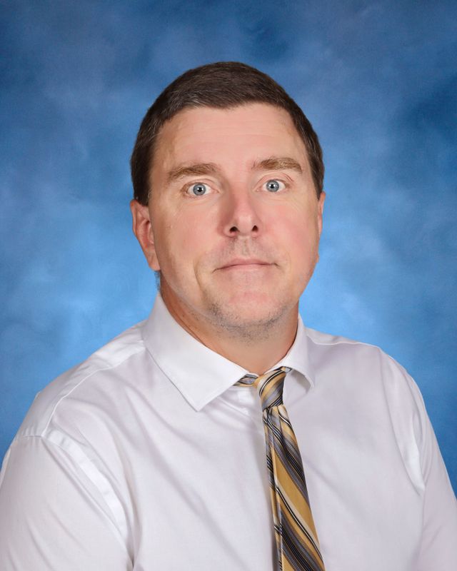 A man with short brown hair in a white shirt and striped tie poses, representing St. Paul's School in front of a blue studio backdrop.
