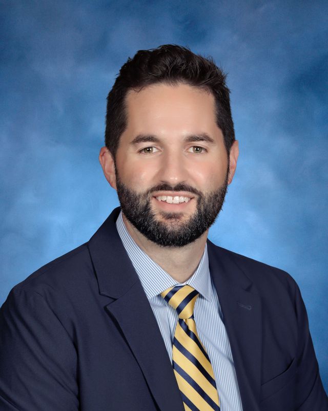 A man with dark hair and a beard, in a navy suit and striped tie, poses before a blue background at St. Paul's School.