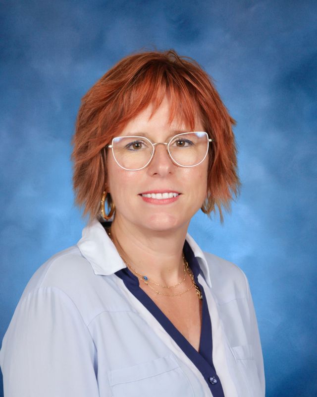 Woman with short red hair and glasses, smiling in front of a blue background, showcasing the welcoming spirit of St. Paul's School.