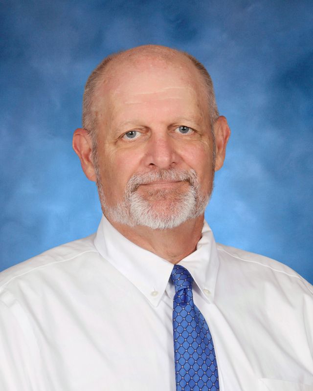 A man with short gray hair and a beard, dressed smartly, poses against a blue background at St. Paul's School on the Northshore.
