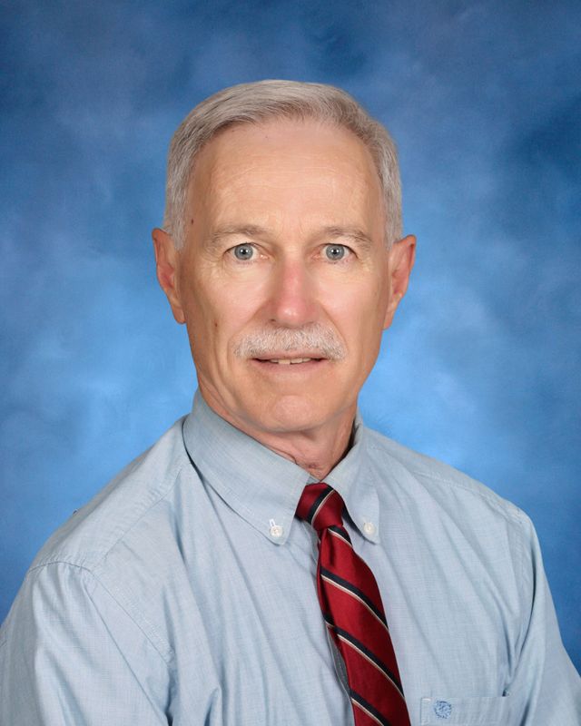 An older man with gray hair and a mustache poses in front of a blue background at St. Paul's School, a top school for boys on the Northshore.