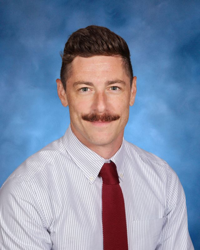 A man with short brown hair and a mustache, in a striped shirt and red tie, poses before a blue backdrop at St. Paul's School.