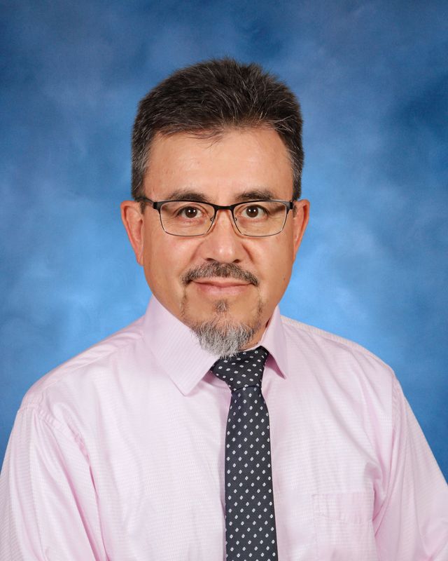 A middle-aged man with glasses and a goatee, in a pink shirt and polka-dot tie, poses before a blue background at St. Paul's School.