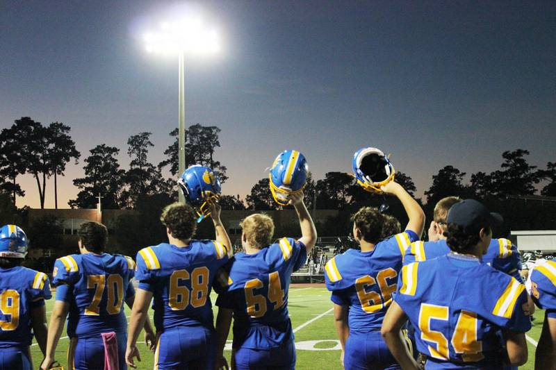 High school football players from St. Paul's School in blue and yellow uniforms stand on the field under stadium lights at dusk.