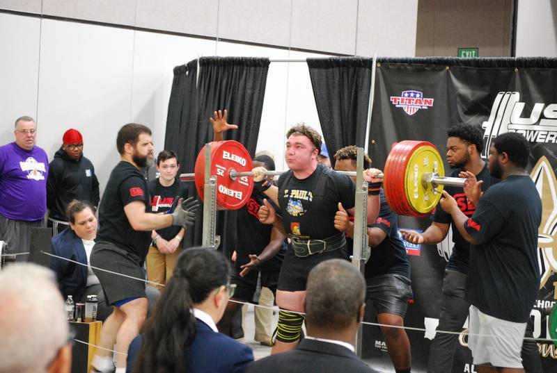 A St. Paul’s School powerlifter prepares to squat a heavily loaded barbell with spotters and judges nearby at a competition.