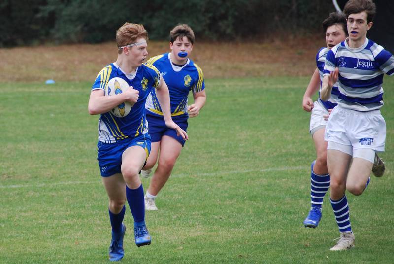 A St. Paul's School rugby player in blue and yellow runs with the ball as teammates and opponents chase on the grassy field.