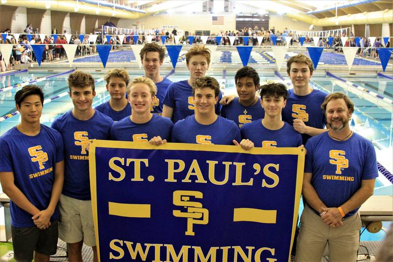 A group of boys and a coach from St. Paul's School pose by a pool, holding a "St. Paul's Swimming" banner and wearing matching blue shirts.