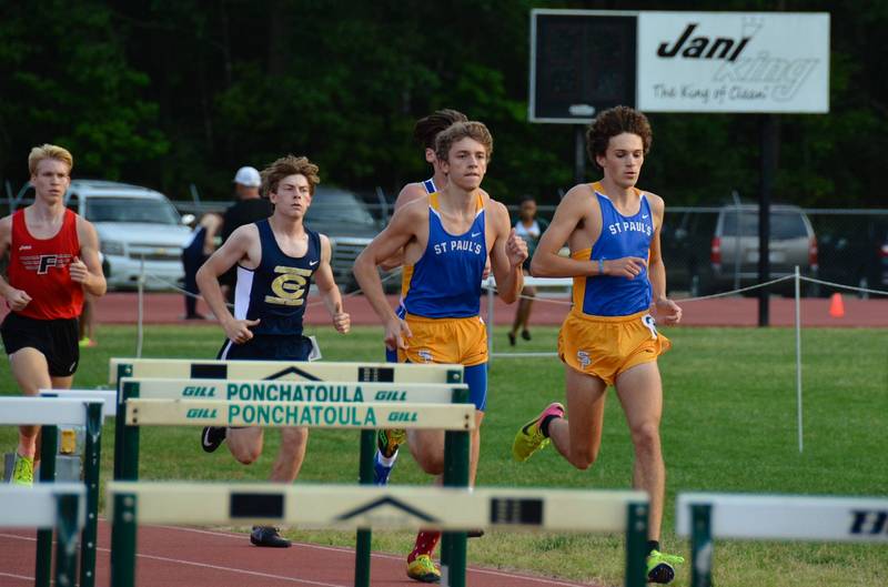 Five high school boys from St. Paul's School race on an outdoor track, hurdles before them and a scoreboard in the background.