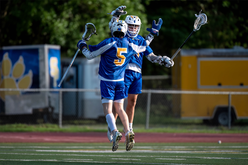 Two St. Paul's School lacrosse players in blue and yellow uniforms jump and celebrate on a field with trailers in the background.