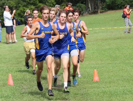 A group of boys in blue and yellow St. Paul’s School uniforms compete in a cross country race on a grassy course.