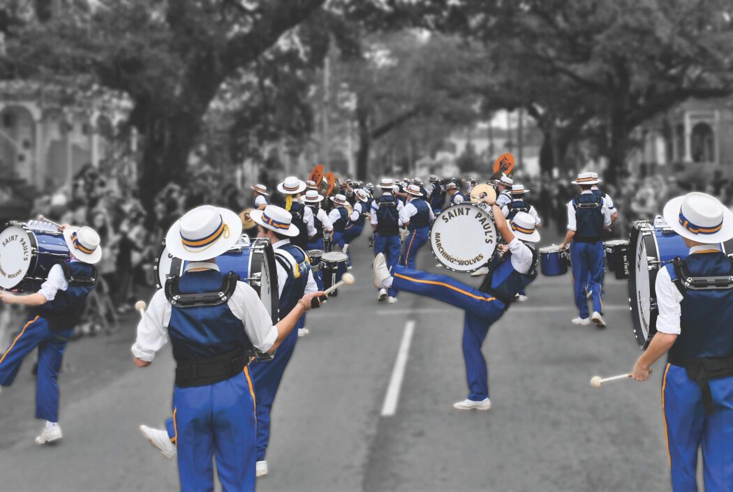 A marching band in blue uniforms and white hats from St. Paul's School performs with drums on a tree-lined street before blurred spectators.