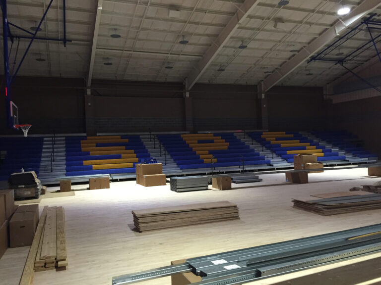 A gymnasium at St. Paul's School with blue and yellow bleachers, a basketball hoop, and construction materials scattered on the floor.