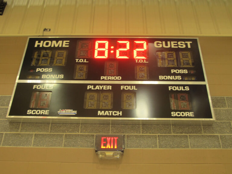 A gymnasium scoreboard at St. Paul’s School shows 8:22 left, with home, guest, fouls, period, scores; an exit sign is below.