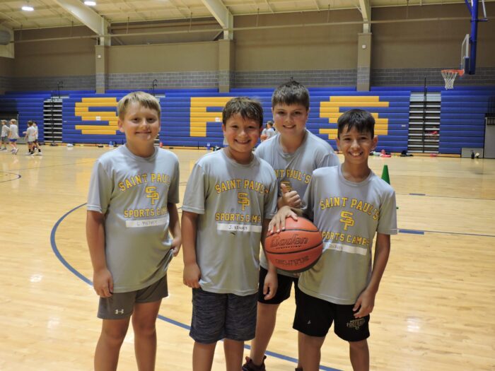 Four boys in St. Paul’s School shirts stand on a basketball court at this top school for boys Northshore LA, with bleachers in the background.