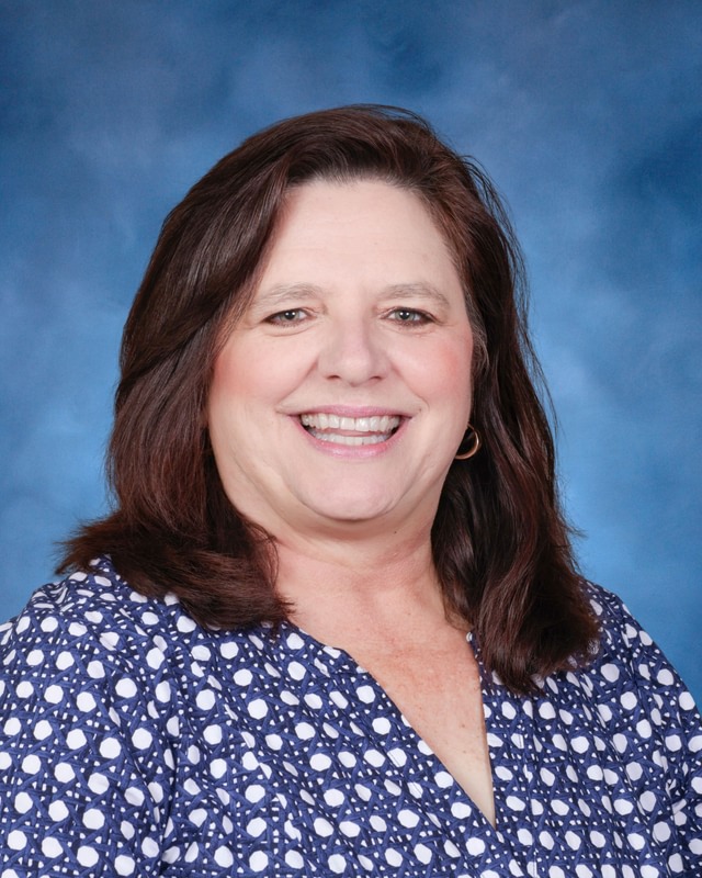 Woman with shoulder-length brown hair wearing a blue and white patterned blouse, smiling in front of a blue background.