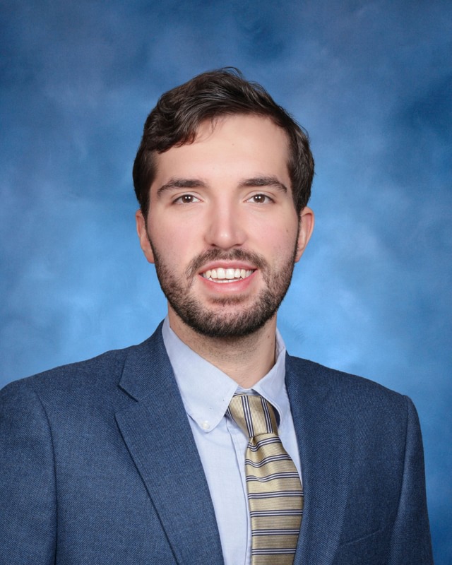 A man with short brown hair and a beard, wearing a blue suit, collared shirt, and striped tie, poses in front of a blue background.