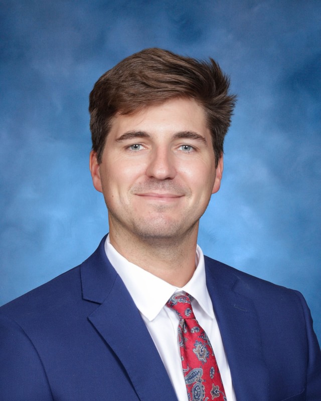A man wearing a blue suit, white shirt, and red patterned tie poses in front of a blue background.