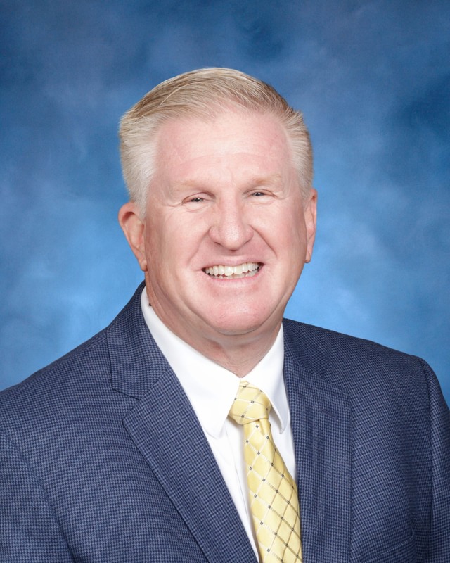 A man in a blue suit, white shirt, and yellow patterned tie smiles in front of a blue studio backdrop.