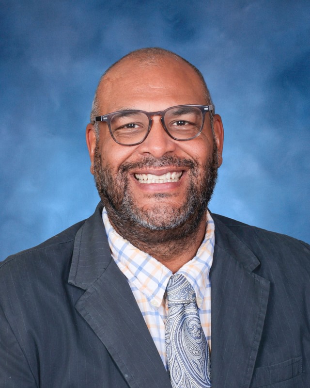 A man with glasses, a beard, and a suit jacket smiles at the camera against a blue studio backdrop.