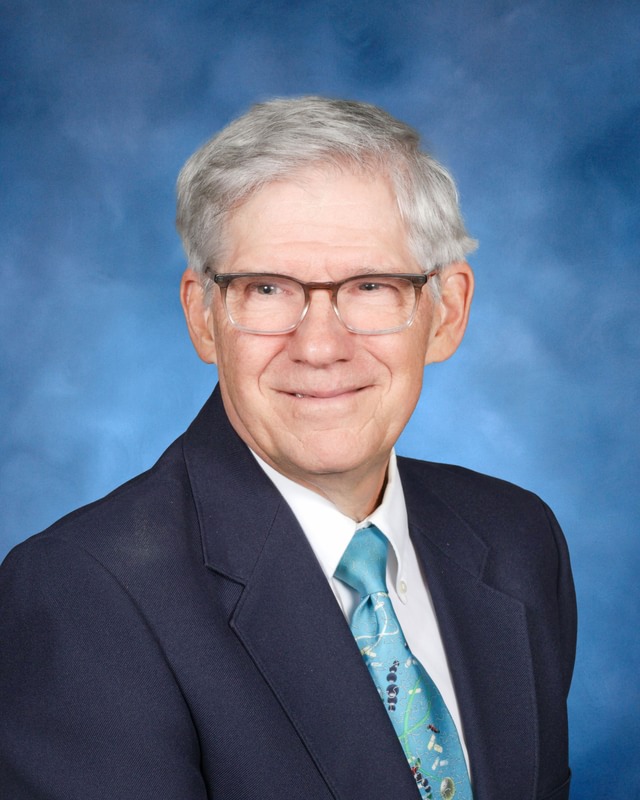 An older man with gray hair and glasses, wearing a dark suit jacket, white shirt, and blue patterned tie, poses in front of a blue backdrop.