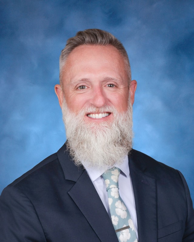 A man with a gray beard and short hair wearing a suit, dress shirt, and floral tie, posing against a blue background.