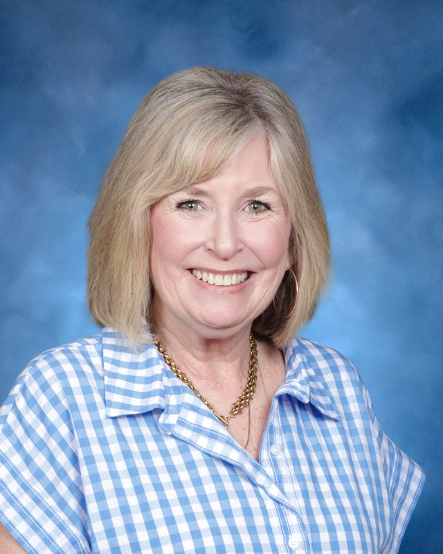 A woman with blonde hair wearing a blue and white checkered shirt and a gold necklace smiles in front of a blue background.