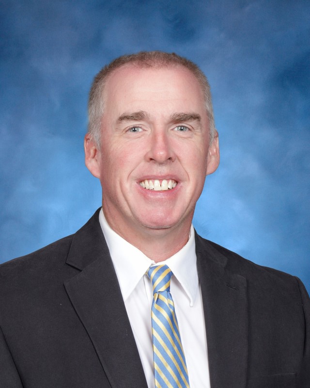 A man in a suit and striped tie smiles in front of a blue studio background.