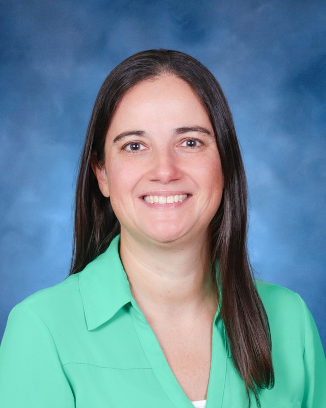 A woman with straight brown hair, wearing a green blouse, smiles at the camera against a blue background.