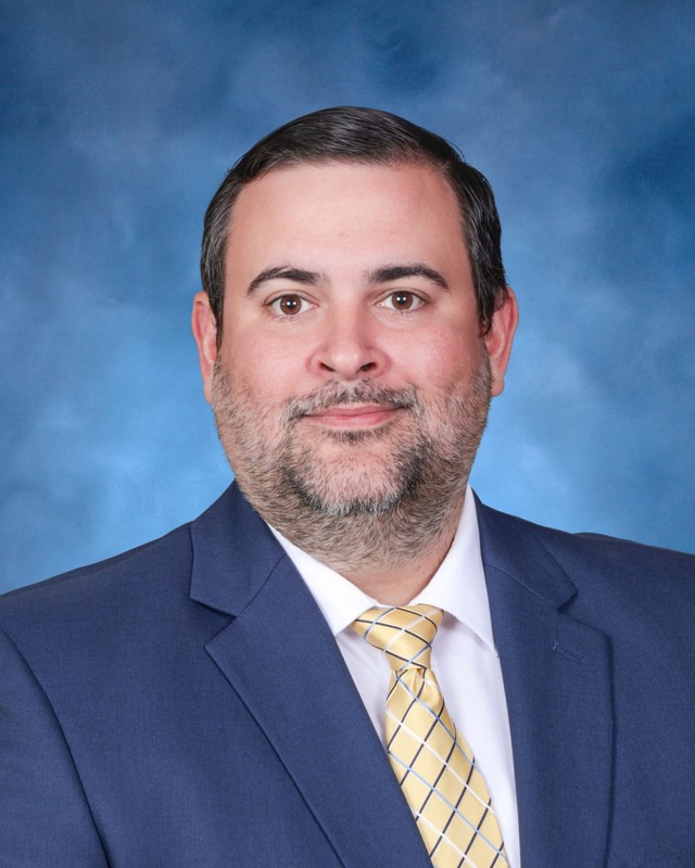 A man in a blue suit and yellow striped tie poses for a formal portrait against a blue background.