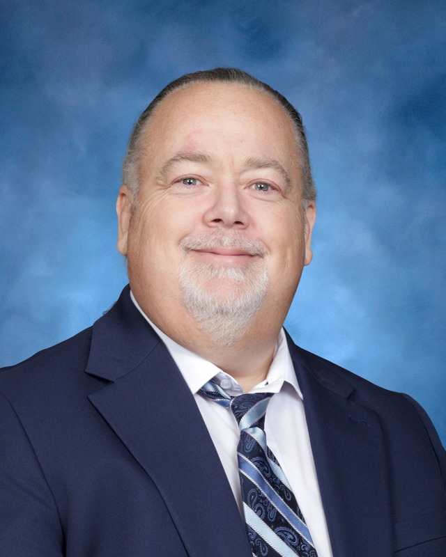 A middle-aged man in a navy suit, white shirt, and patterned tie poses in front of a blue studio background, smiling slightly.