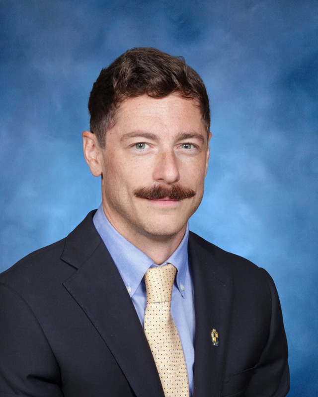 A man with short brown hair and a mustache wearing a suit, blue shirt, and yellow tie poses in front of a blue backdrop.
