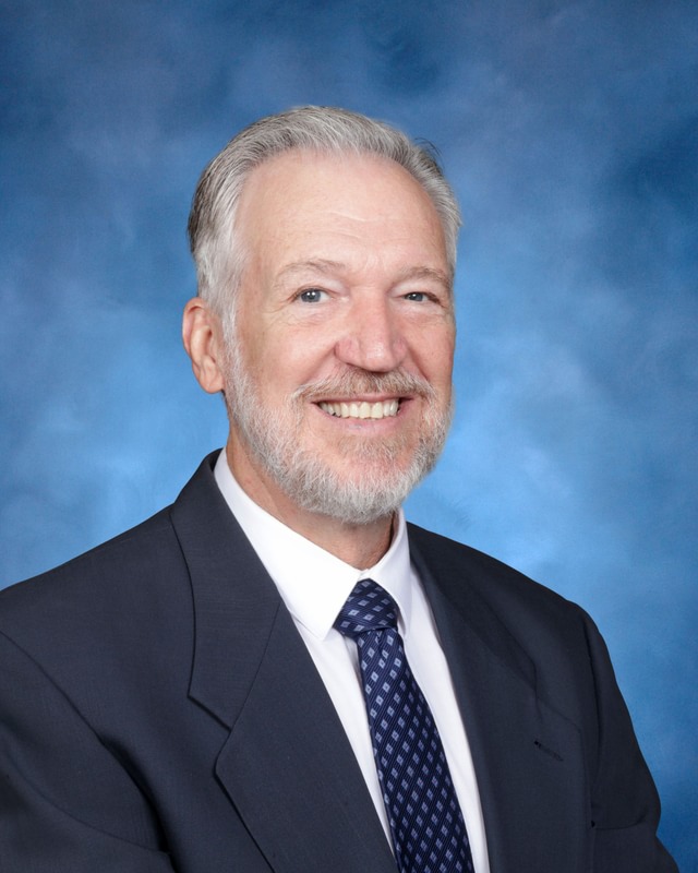 A man with gray hair and beard in a suit and tie smiles in front of a blue studio background.