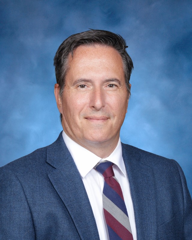 A man in a blue suit, white shirt, and striped tie poses against a blue studio background, looking at the camera and smiling slightly.