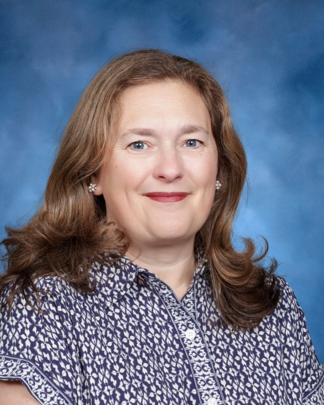 A woman with long brown hair smiles at the camera, wearing a patterned blouse, against a blue studio background.