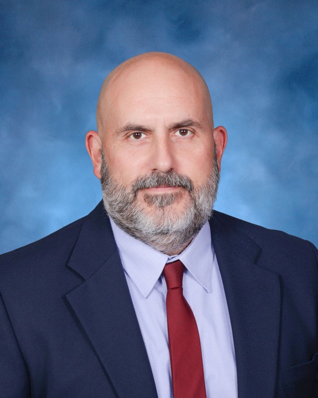 A man with a bald head and gray beard wearing a dark suit, light blue shirt, and red tie poses in front of a blue background.