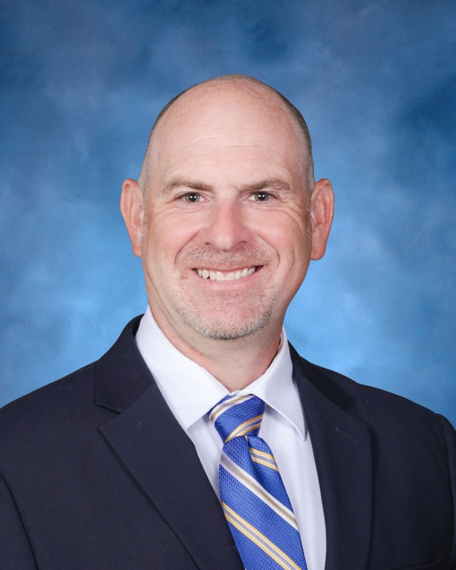 A man in a dark suit and blue striped tie smiles at the camera against a blue studio background.