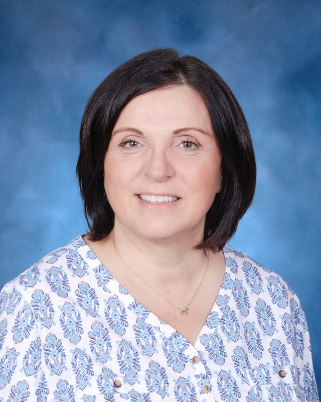 A woman with straight dark hair, wearing a white shirt with a blue floral pattern, poses in front of a blue background.