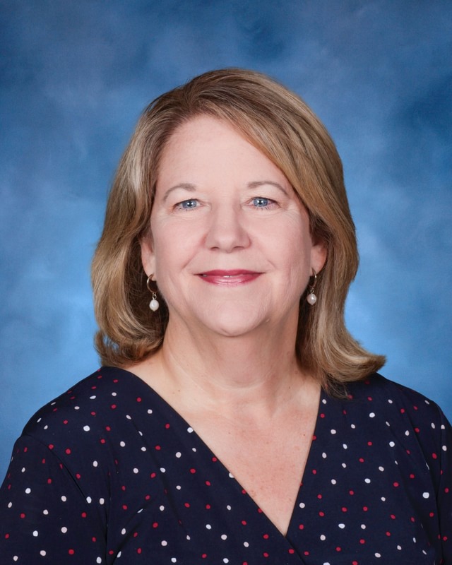 A middle-aged woman with shoulder-length blonde hair, wearing a dark polka dot blouse, smiles in front of a blue studio background.