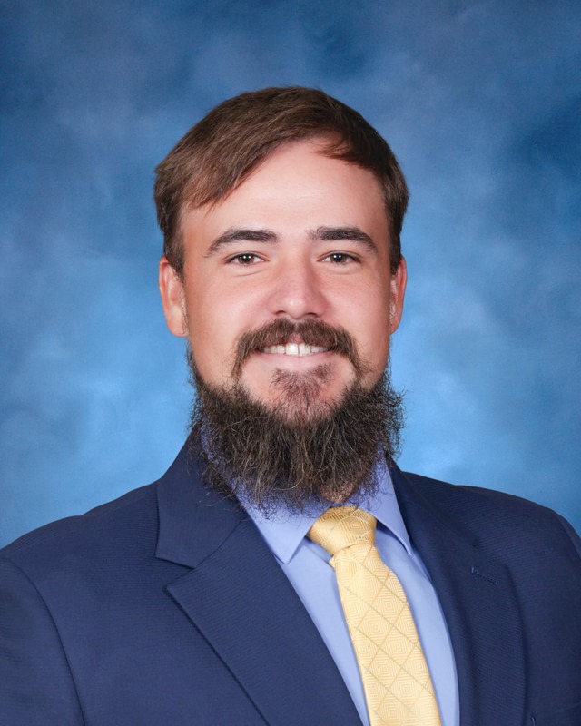 Man with brown hair and beard wearing a blue suit, light blue shirt, and yellow tie, posing in front of a blue background.