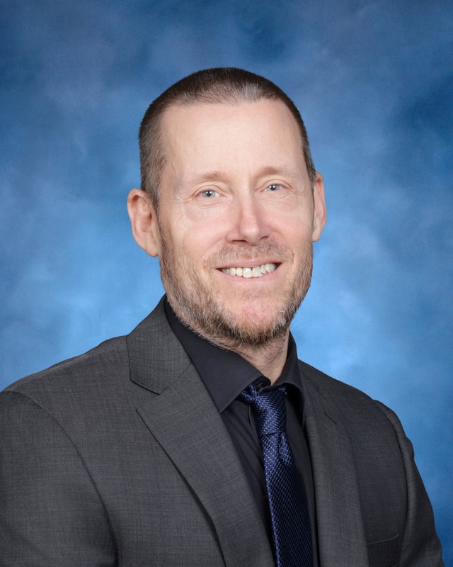 A man with short hair and a beard wearing a dark suit, black shirt, and blue tie, smiling in front of a blue background.