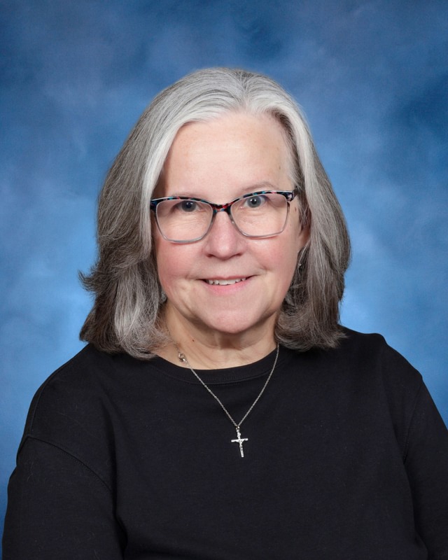 A woman with gray hair, glasses, and a black top, wearing a cross necklace, poses in front of a blue studio background.