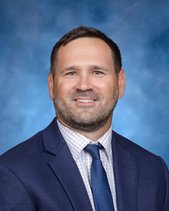 A man with short brown hair and a beard wearing a blue suit, checkered shirt, and blue tie against a blue background.