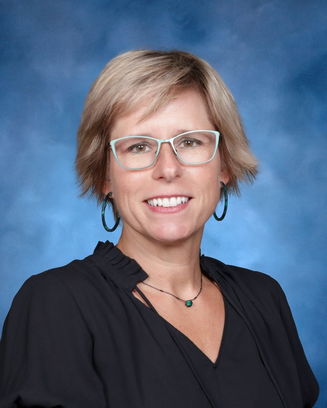 A woman with short blonde hair, glasses, and hoop earrings smiles in front of a blue background. She is wearing a black blouse.