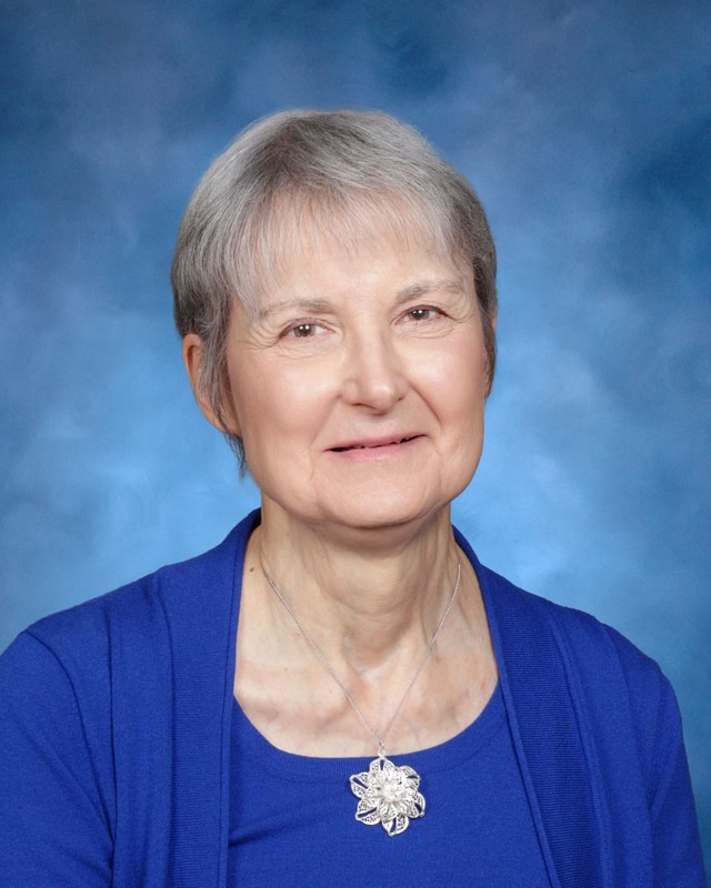 An older woman with short gray hair wears a blue top and a floral pendant necklace, posing in front of a blue background.