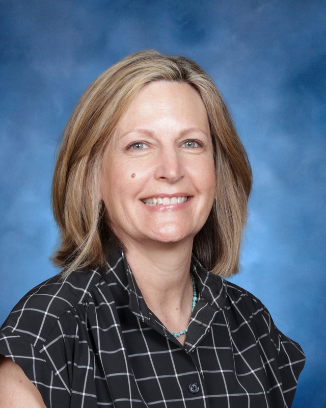 A woman with shoulder-length blonde hair, wearing a black and white plaid shirt, smiles in front of a blue background.