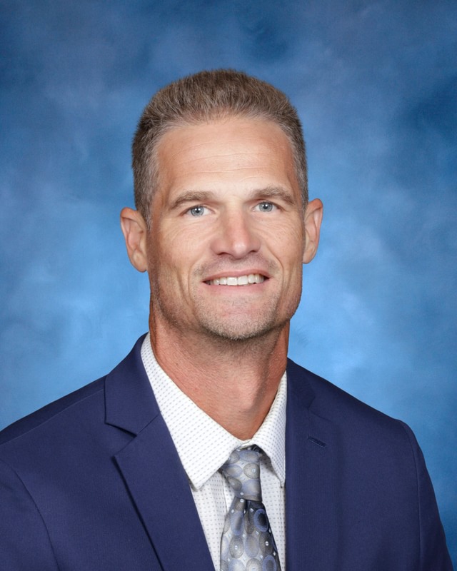 A man in a blue suit, white shirt, and patterned tie poses for a formal portrait against a blue studio background.