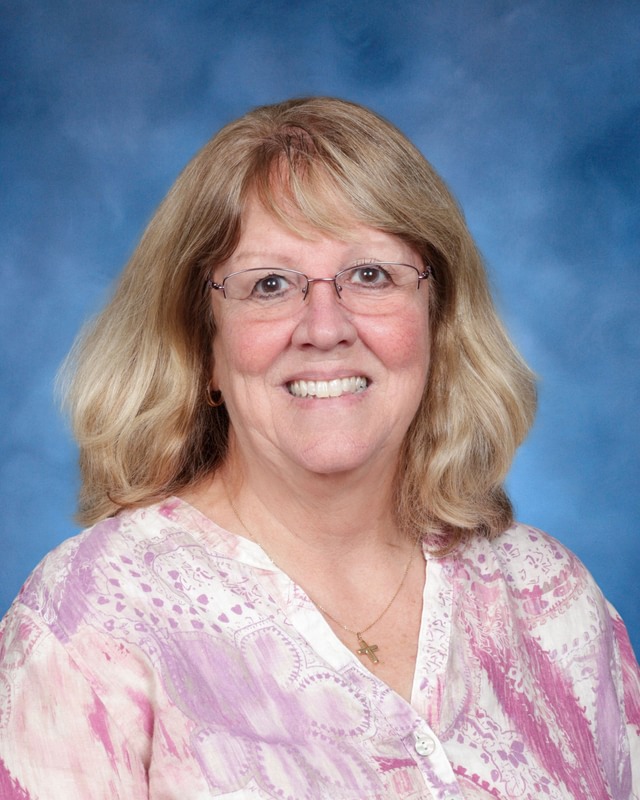 A woman with shoulder-length blonde hair, glasses, and a pink patterned blouse smiles in front of a blue background.