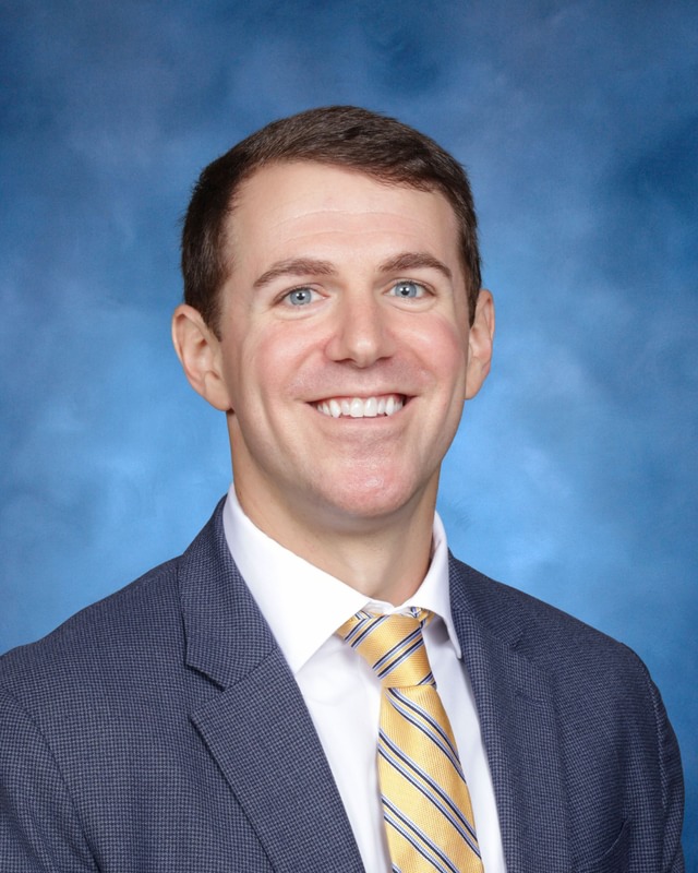 A man in a suit, white shirt, and yellow striped tie smiles at the camera against a blue background.