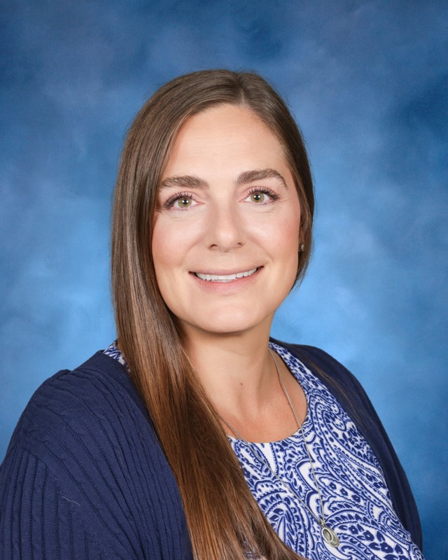 A woman with long brown hair, wearing a blue patterned top and navy cardigan, poses in front of a blue background, smiling at the camera.